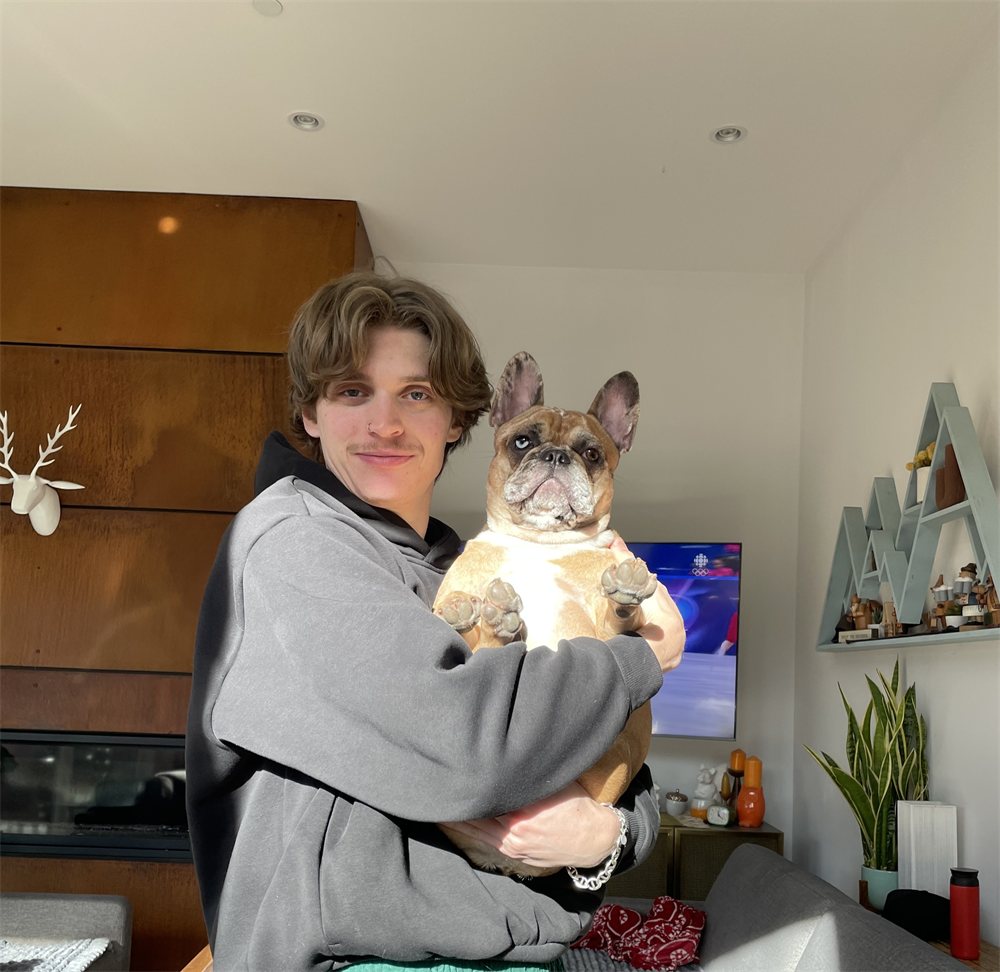 Binx smiling in a grey hoodie, holding a thoroughly unimpressed French bulldog in a sunlit living room